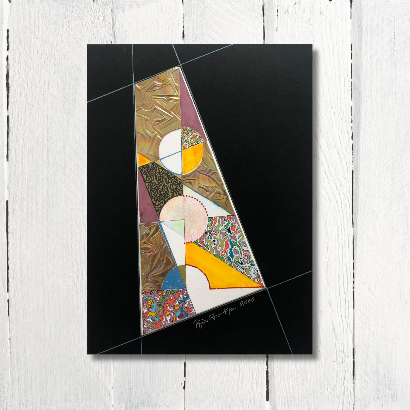 A geometric collage art piece with a combination of drawings, engraved bronze, and a black cardboard frame, placed on a white wooden surface.