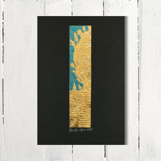 An engraving of trees among rocks, made on a mixed metal foil with turquoise details on black cardboard, displayed against a white wooden background.